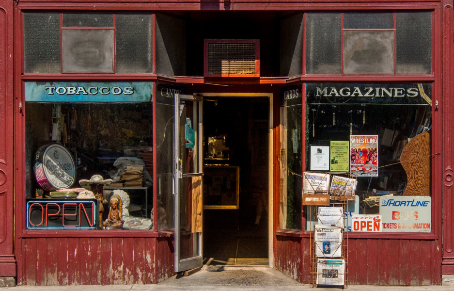 Ben Asen Personal Work Photo: Color photo of a red general store in Owego New York with a neon light Open sign with tobaccos and magazines signs in the window and newspapers for sale outside the store.