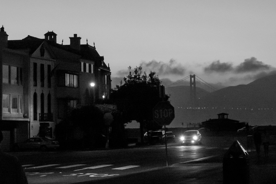 Ben Asen Personal Work Photo: Black and white photo of a street at night with oncoming car with lights with the Golden Gate Bridge in the background.